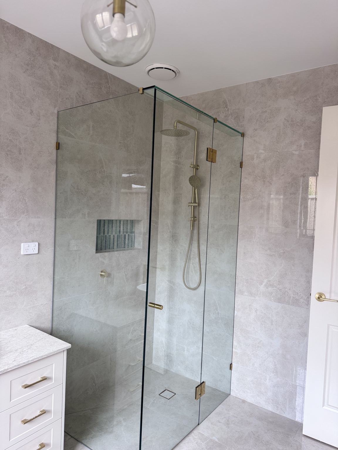 Clear frameless corner shower with gold-toned hardware in a pale tiled bathroom