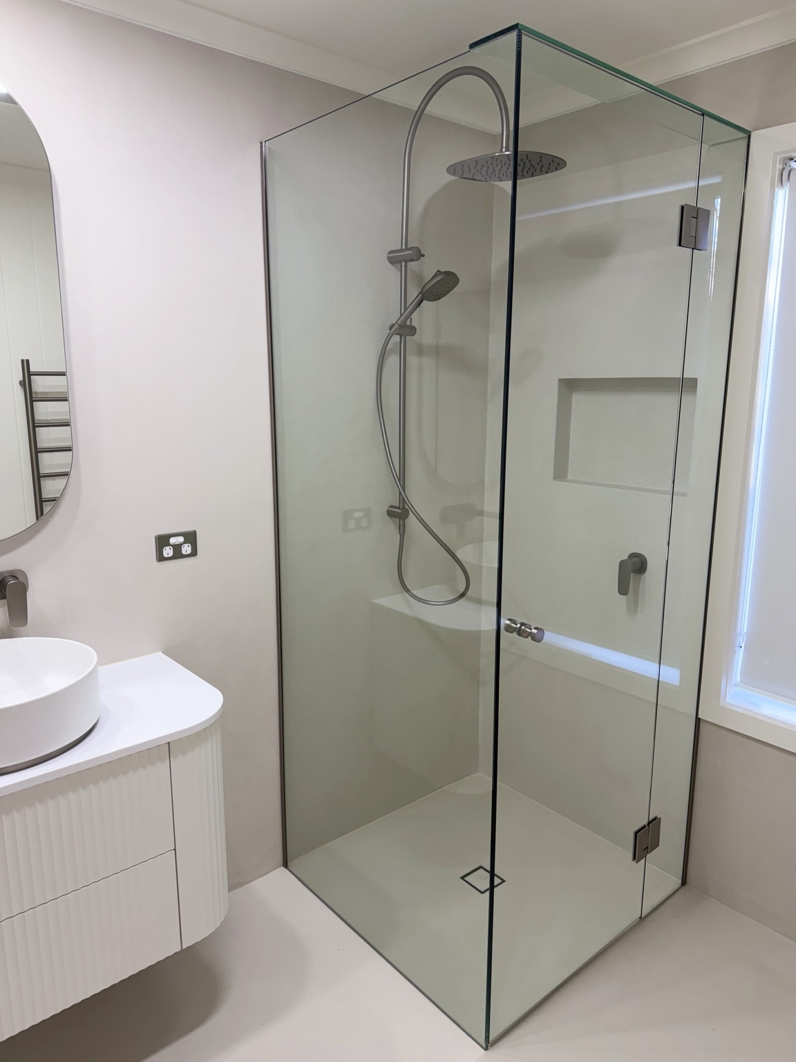 Clear frameless corner shower beside a curved white vanity